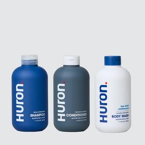 Three Huron shower bottles: blue shampoo, gray conditioner, white Sea Salt body wash on pale gray background #scent_sea salt + driftwood