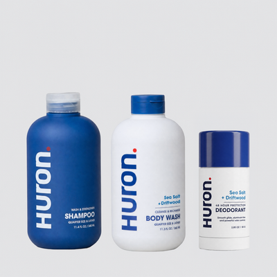 Three Huron personal care products on a light gray background: a blue shampoo bottle, a white body wash bottle, and a white deodorant stick with blue cap.