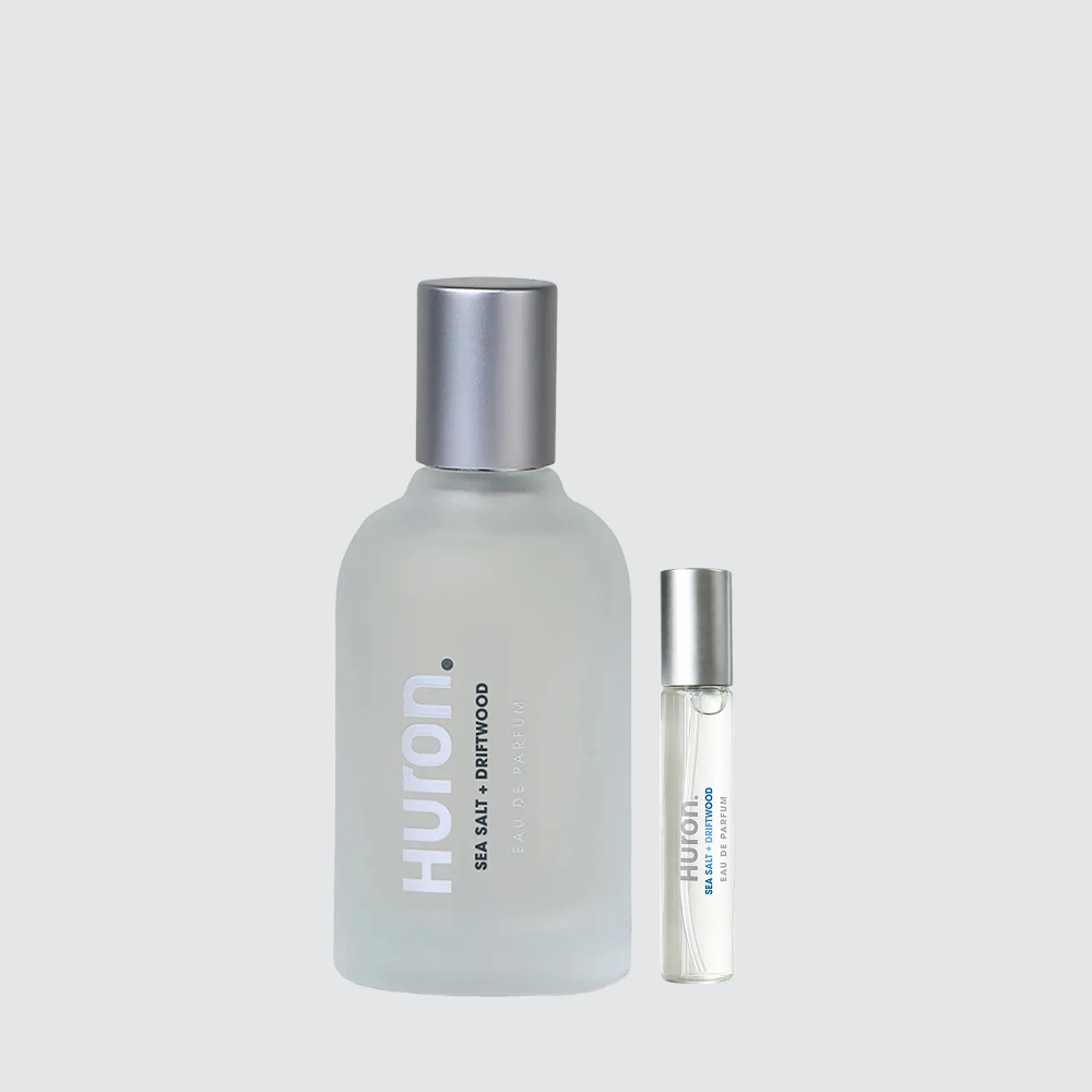 HURON cologne: frosted glass bottle and travel spray labeled HURON and SEA + DRIFTWOOD on pale gray background
