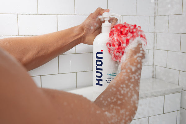 Arm pressing pump of HURON bottle labeled 'Original Citrus + Eucalyptus CLEANSE & RECOVER BODY WASH' with soapy red pouf