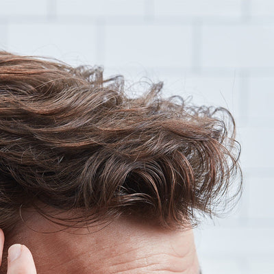 Close-up of a man's brown wavy hair styled with textured lift at the front; hand near ear showing hold.
