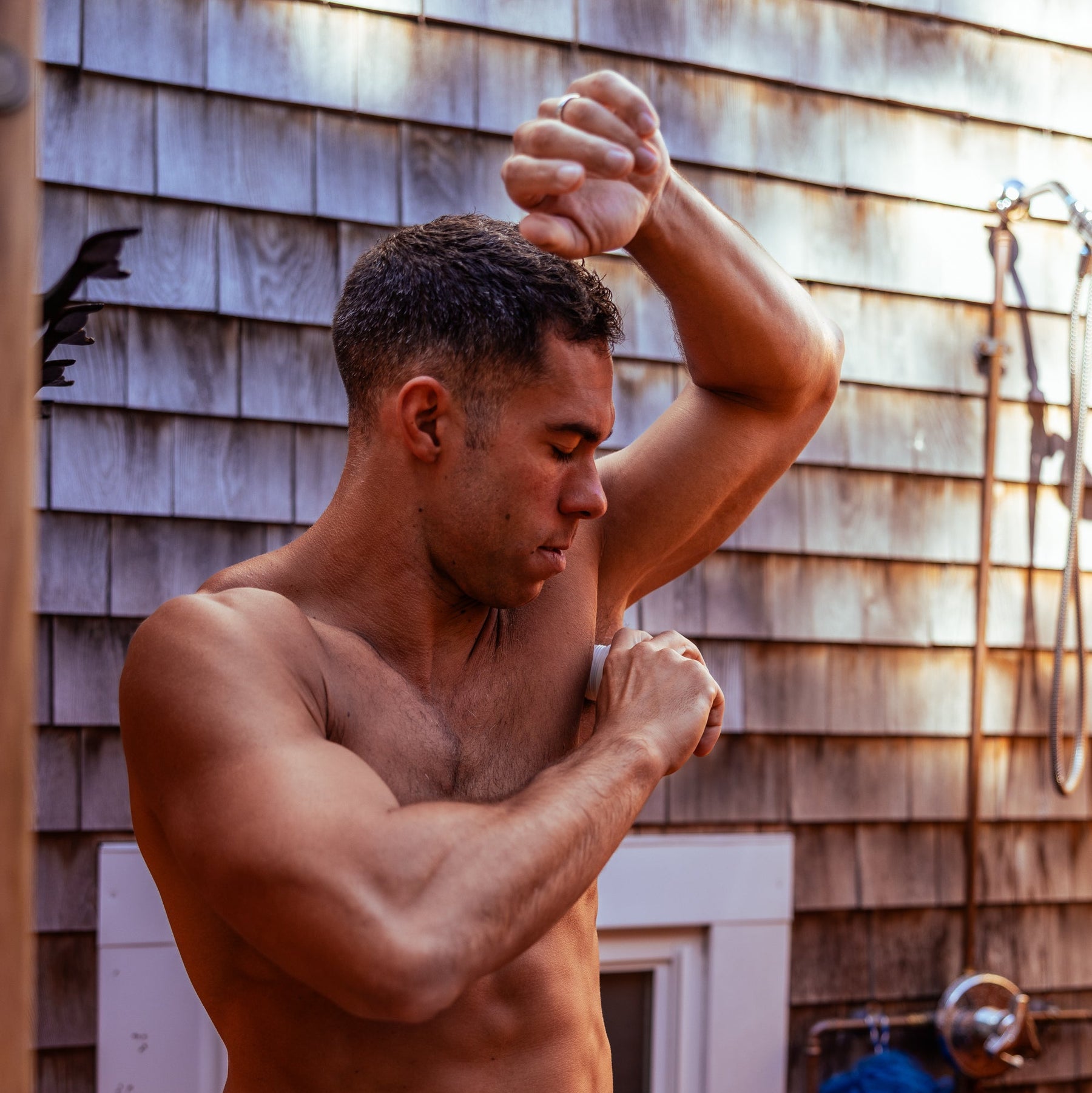 Shirtless man applying deodorant to his underarm outside next to a wooden shingled wall