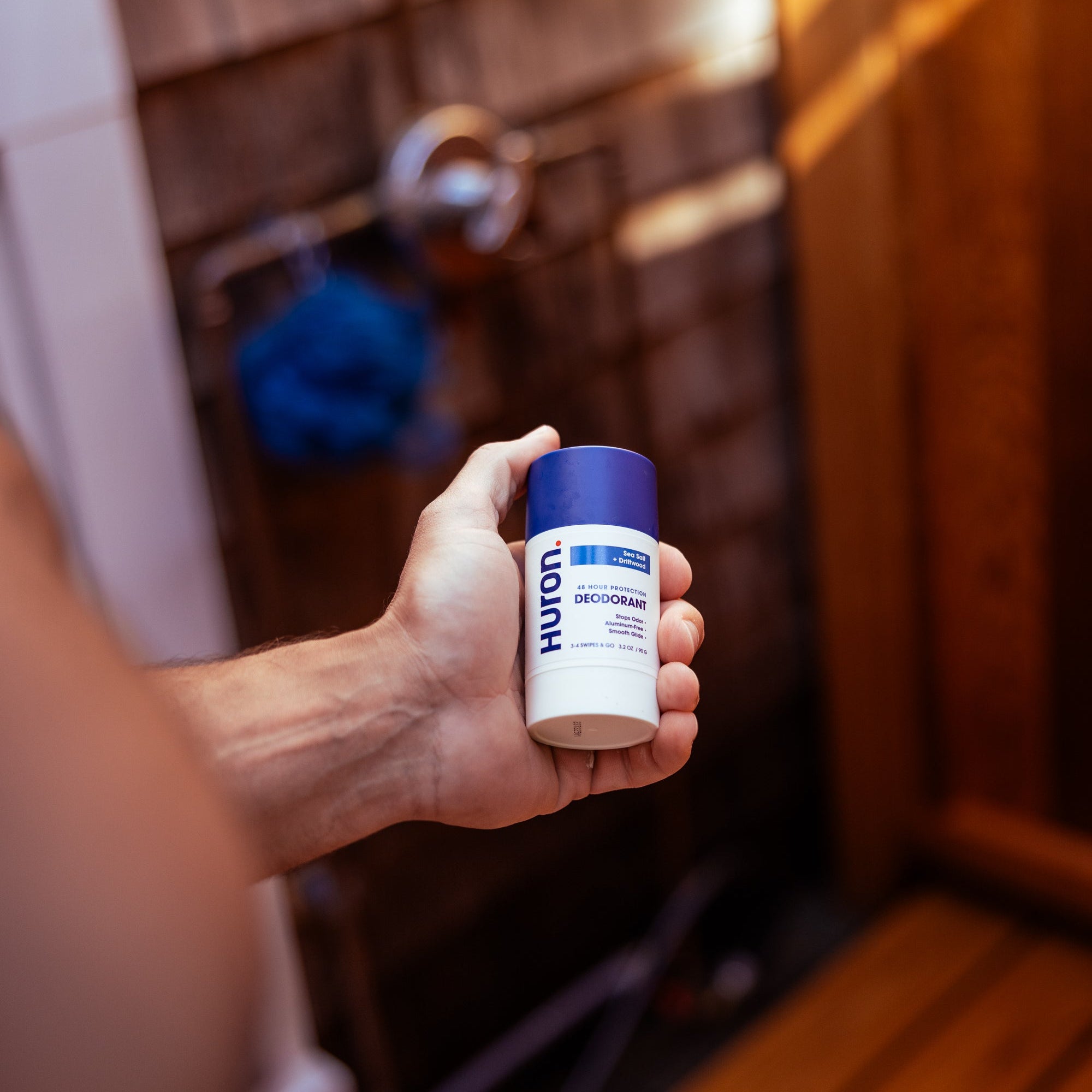 Hand holding Huron Deodorant stick with blue cap in a wooden-tiled shower, blurred showerhead in background.