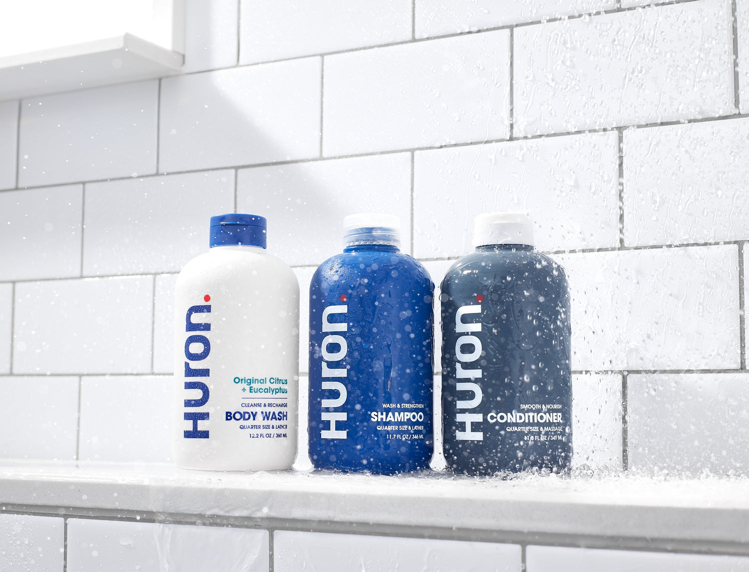 Three Huron shower bottles on a wet tile shelf: Body Wash (Original Citrus + Eucalyptus), Shampoo, Conditioner.