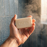 Close-up of Huron bar soap held in a hand showing the embossed 'Huron' logo, wet under running shower water with steam and tiled background