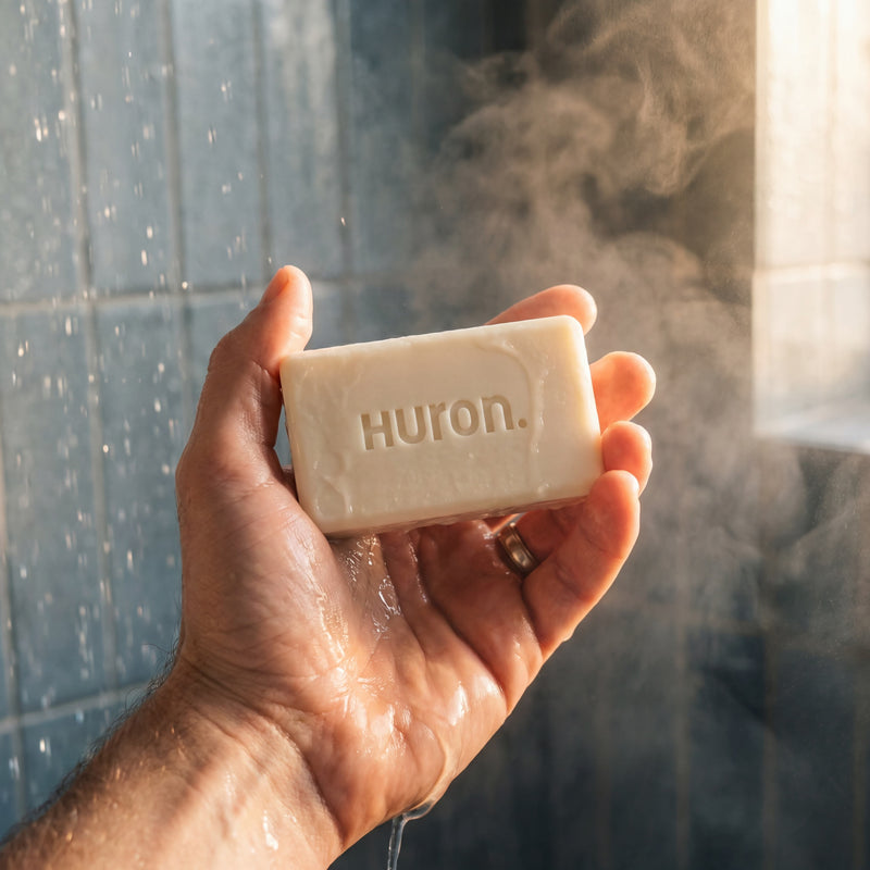Close-up of Huron bar soap held in a hand showing the embossed 'Huron' logo, wet under running shower water with steam and tiled background