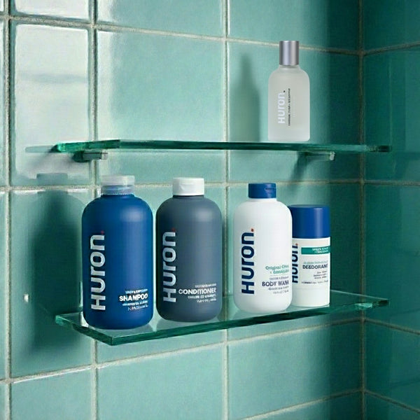 Glass shower shelf holding HURON bottles labeled SHAMPOO, CONDITIONER, BODY WASH and DEODORANT against green tiles.