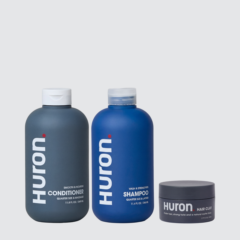 Front view of Huron shampoo, conditioner, and hair clay in blue and gray packaging on a light gray background