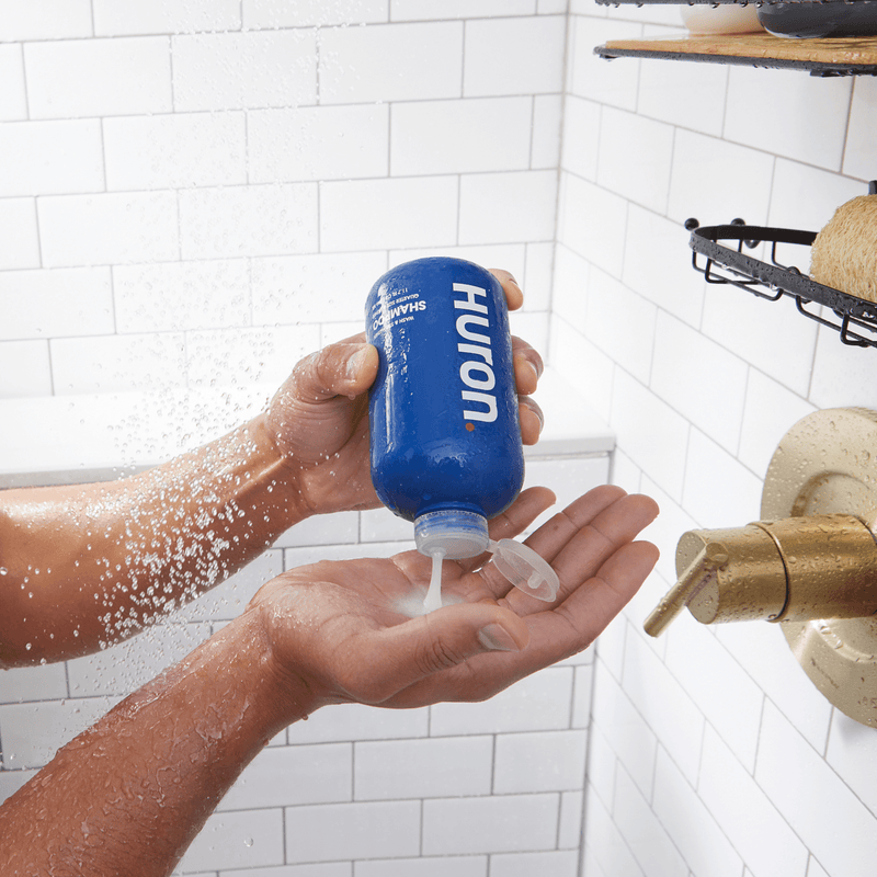 Person in shower squeezing Huron SHAMPOO bottle, white lather dispensed into cupped hand