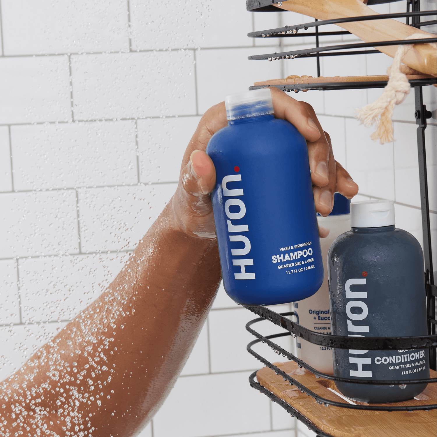 Hand holding a blue bottle of Huron Shampoo in a shower with a black wire rack holding other Huron hair care products.