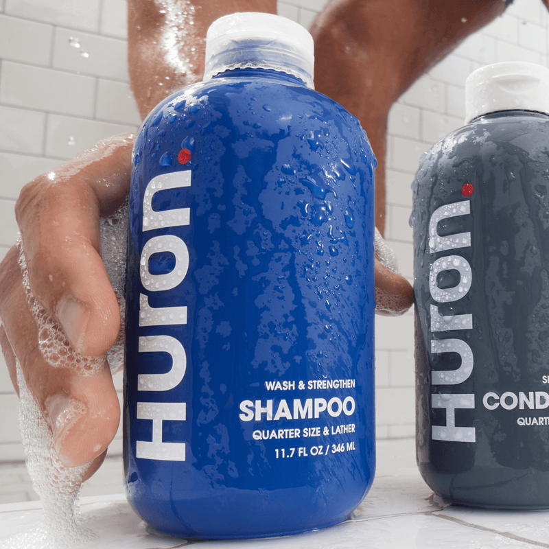 Close-up of blue Huron Wash and Strengthen shampoo bottle held by soapy hand, wet bottle and visible SHAMPOO 11.7 fl oz