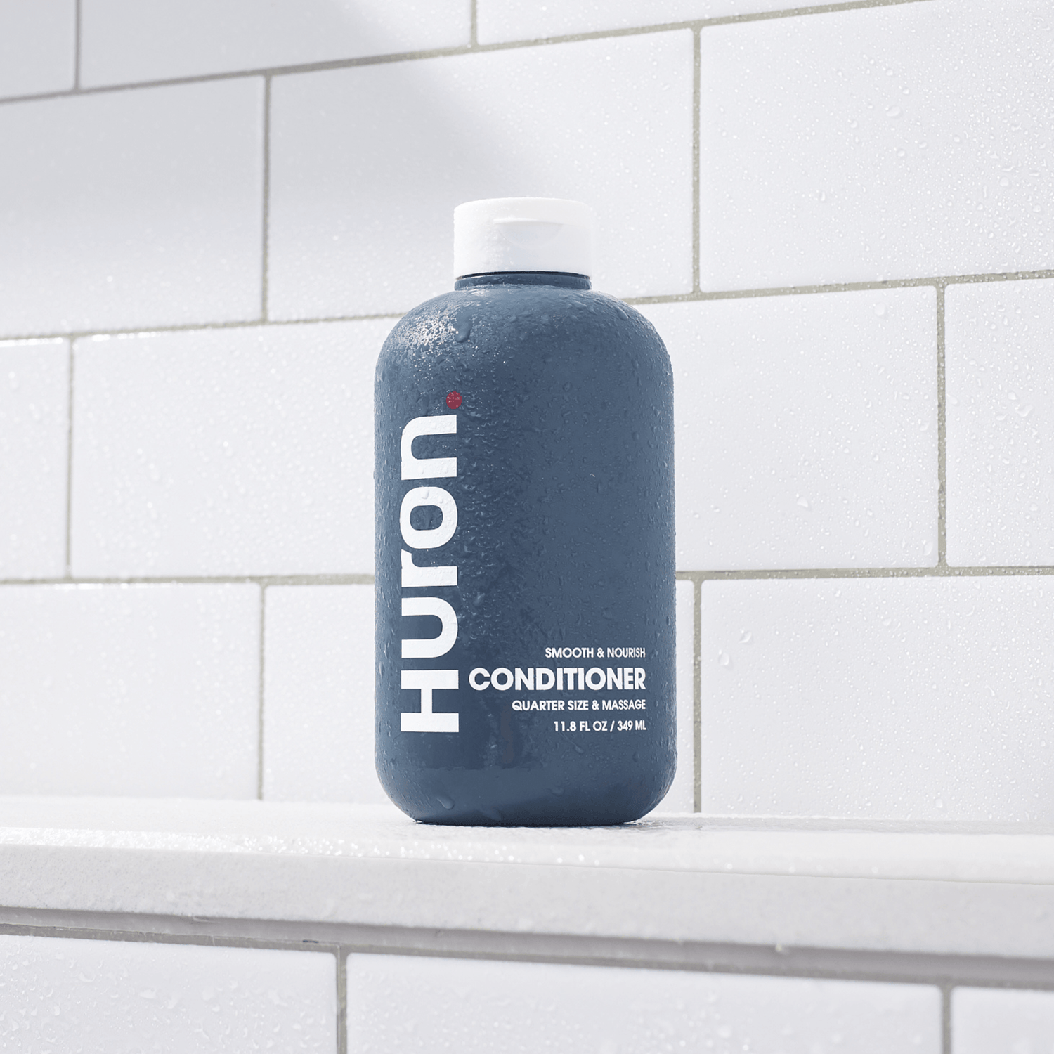 Blue bottle on tile with text: HURON SMOOTH & NOURISH CONDITIONER QUARTER SIZE & MASSAGE 11.8 FL OZ / 349 ML