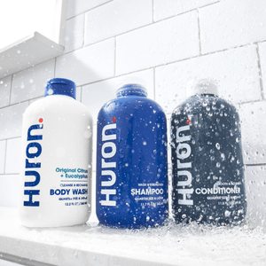 Front view of three Huron bottles, Body Wash, Shampoo, and Conditioner, with water splashing in a tiled shower showcasing Huron branding