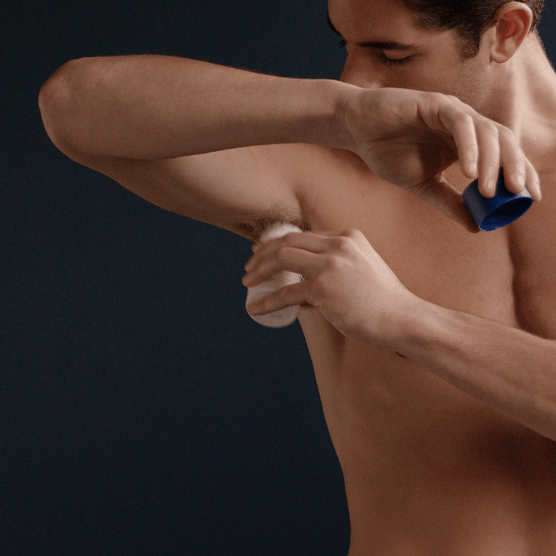Man applying roll-on deodorant to underarm, holding white deodorant stick and blue cap.